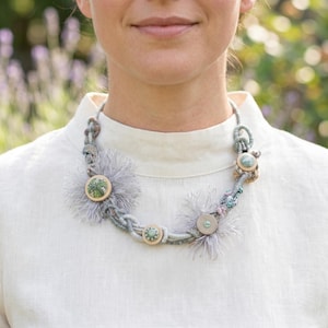 Op de afbeelding: Een statement ketting met gevlochten lichtgrijs koord, zachte grijze franjeaccenten en diverse ronde houten knopen met groen-witte patronen. De ketting wordt om de hals gedragen en toont een gelaagd, gestructureerd ontwerp met gemengde materialen.