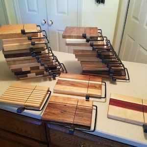 Peut inclure: Une collection de planches à fromage en bois avec des poignées en métal. Les planches sont faites de différents types de bois, dont l'érable, le noyer et le cerisier. Les planches sont empilées en un tas, et certaines sont posées à plat sur un comptoir.