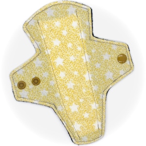 Puede incluir: Una compresa menstrual de tela reutilizable amarilla y blanca con un patrón de estrellas y ribete negro. La compresa tiene tres broches dorados en un lado y un broche dorado en el otro.