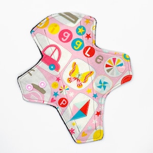 Puede incluir: Una compresa de tela rosa con un estampado colorido de letras, estrellas, mariposas y un coche. La compresa tiene dos broches de presión rosas en el lateral.