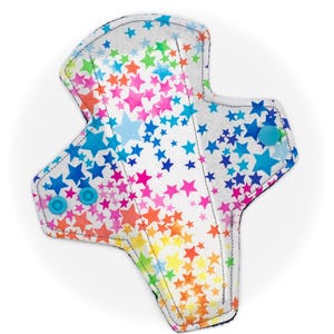 Puede incluir: Una compresa menstrual de tela reutilizable con fondo blanco y un patrón de estrellas de colores. La compresa tiene dos broches azules en el lateral.