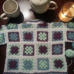 May include: A crocheted cardigan with a square pattern in shades of green, blue, and purple. The cardigan is made with white yarn and has a simple, classic design.