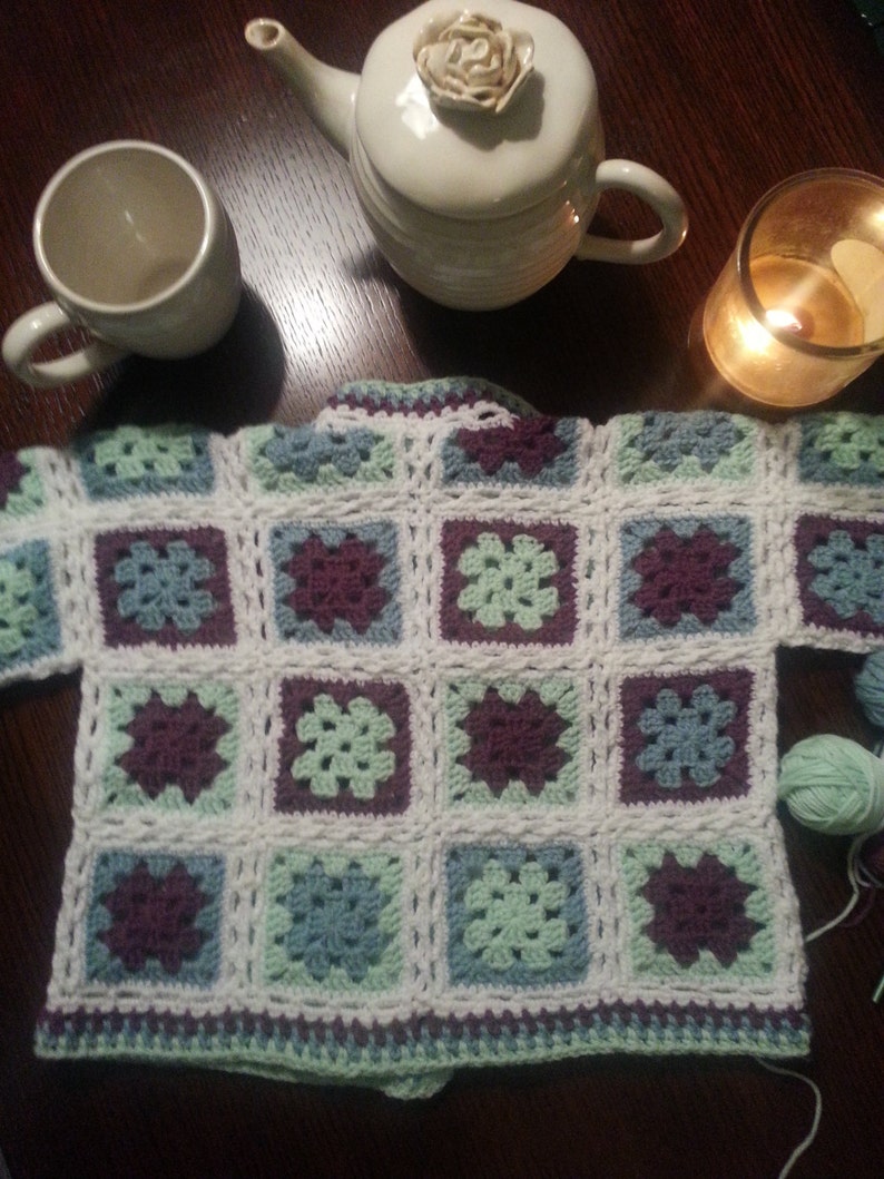 May include: A crocheted cardigan with a square pattern in shades of green, blue, and purple. The cardigan is made with white yarn and has a simple, classic design.