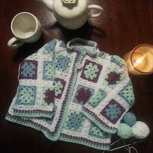 May include: A crocheted cardigan with a granny square pattern in shades of white, green, and purple. The cardigan has a button closure and is made with a soft yarn.