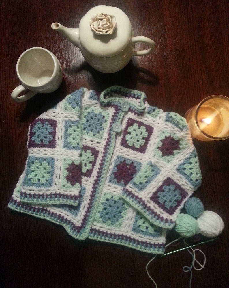May include: A crocheted cardigan with a granny square pattern in shades of white, green, and purple. The cardigan has a button closure and is made with a soft yarn.