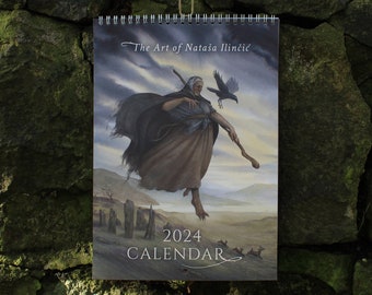 May include: A 2024 calendar featuring artwork by Nataša Ilinčić, depicting a woman with a raven in a fantasy landscape. The calendar is spiral-bound and hangs from a natural twine string against a stone wall.