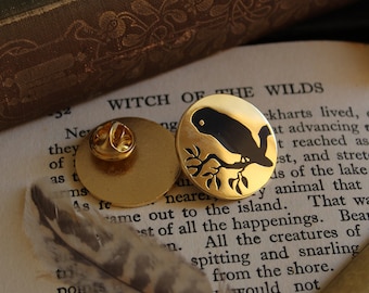 Può includere: Spilla dorata con un uccello nero posato su un ramo. La spilla è rotonda e ha un retro a spillo. L'uccello e il ramo sono neri. La spilla è su un libro con il testo "WITCH OF THE WILDS".
