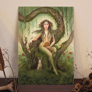 May include: A watercolor painting depicts a woman with long brown hair adorned with a floral crown, seated on a tree branch. She holds a golden symbol. A white rabbit sits below. The artwork features green foliage and red roses.