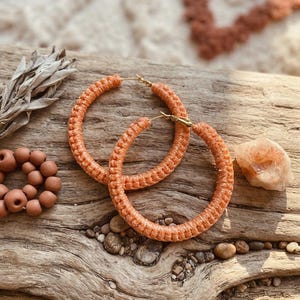 May include: A pair of large hoop earrings wrapped in textured, peach-colored cord. The earrings feature a gold-toned metal post and clasp mechanism. The circular design is consistent in thickness and color, showcasing a woven, knotted surface texture throughout the entire circumference of each hoop.