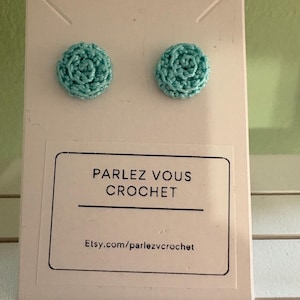 Peut inclure: Une paire de boucles d'oreilles en crochet turquoise, présentées sur une carte blanche. Les boucles d'oreilles rondes ont un motif texturé en forme de fleur. La carte affiche le texte "PARLEZ VOUS CROCHET" et une adresse de site web.