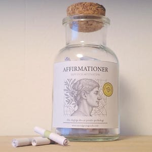 May include: A clear glass jar with a cork stopper, labelled "Affirmationer." The label features a classical bust and floral elements. Rolled paper scrolls are scattered in front of the jar, suggesting motivational content.