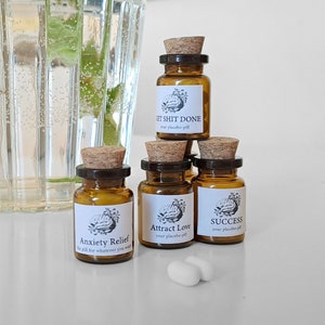 Può includere: Cinque piccoli flaconi di vetro marrone con tappi di sughero ed etichette bianche. Le etichette hanno testo nero e un piccolo grafico di un cervello. Il testo sulle etichette recita "Anxiety Relief", "Get Shit Done", "Attract Love", "Success" e "Your Placebo Pill". Due pillole bianche sono davanti ai flaconi.