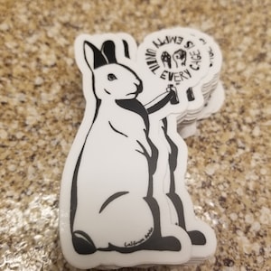 May include: A stack of white stickers featuring a black and white illustration of a rabbit holding a spray can. The text on the sticker reads "Until Every Cage Is Empty". The stickers are on a speckled brown surface.