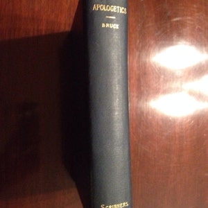 May include: A dark blue book titled "Apologetics" by Bruce, with "Scribners" printed at the bottom. The text is in gold lettering. The book is standing upright on a dark wooden surface.