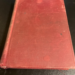 Puede incluir: Un libro antiguo, de color rojo, con una cubierta texturizada y signos de desgaste. La cubierta presenta texto descolorido y pequeños daños. Los bordes del libro están ligeramente deshilachados. La apariencia general sugiere un artículo antiguo.