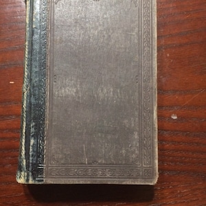 Puede incluir: Un libro antiguo con una cubierta gris desgastada y un borde decorativo. El lomo es azul oscuro y muestra signos de desgaste. El libro está cerrado y apoyado sobre una superficie de madera.