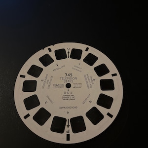 May include: A white View-Master reel with the text "745 TELEVISION STARS" and names including Ed Sullivan and Groucho Marx. The circular reel has rectangular viewing windows and directional arrows, a classic entertainment item.