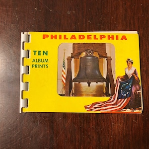 May include: A vintage Philadelphia souvenir album with a yellow cover. The cover features the text "PHILADELPHIA" and "TEN ALBUM PRINTS." Inside the album's window is an image of the Liberty Bell and a woman holding a flag.
