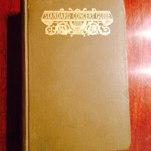 Puede incluir: Un libro antiguo titulado "Standard Concert Guide" con letras doradas y elementos decorativos en una cubierta verde apagada. El libro es rectangular y parece estar en buen estado, descansando sobre una superficie de madera oscura.
