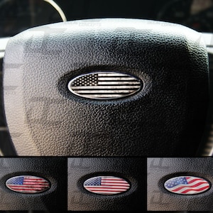 May include: A black steering wheel with a silver oval emblem featuring a distressed American flag design. The emblem is centered on the steering wheel. Below the steering wheel are three smaller images of the same emblem with different distressed flag designs: one with red, white, and blue colors, one with red and white colors, and one with blue and white colors.