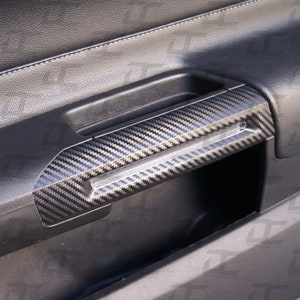 Könnte beinhalten: Ein schwarzer Kohlefaser-Türgriff mit strukturierter Oberfläche. Der Griff ist an einer schwarzen Ledertürverkleidung eines Autos angebracht.