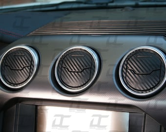 May include: Three black carbon fiber air vent covers for a car dashboard. The covers are round and have a silver trim.