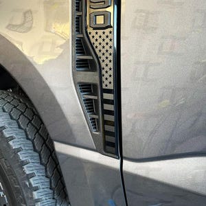 May include: A close-up of a vehicle's side vent with a black and silver American flag design. The vent is part of a gray vehicle, and the tire is visible in the lower left corner. The flag design features stars and stripes.