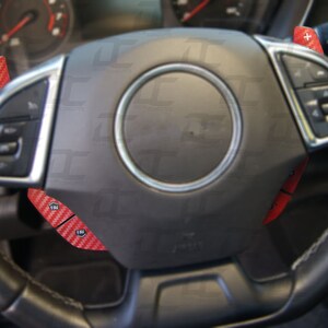 May include: A black leather steering wheel with red carbon fiber paddle shifters. The paddle shifters are labeled with the letters "FM".