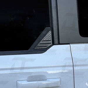 May include: A white truck with a black and gray American flag decal on the rear window. The decal is in the shape of a triangle and is placed on the top corner of the window.