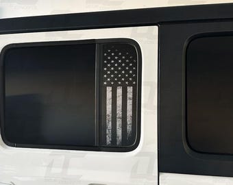 Könnte beinhalten: Ein weißer Jeep Wrangler mit schwarzen Fensterverkleidungen und einem abgenutzten amerikanischen Flaggenaufkleber am hinteren Beifahrerfenster.