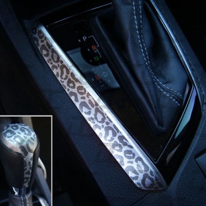 May include: A close-up of a car's gear shift and center console with a leopard print design. The gear shift is silver and has a black leather knob. The console is black and has a silver trim with a leopard print design.