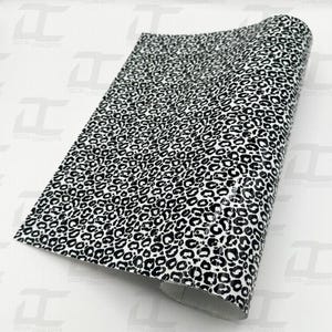 Könnte beinhalten: Ein rechteckiger Bogen aus selbstklebender Vinylfolie mit einem sich wiederholenden schwarzen Leopardenmuster auf weißem Grund. Das Material hat eine glänzende Oberfläche und ist an einem Ende teilweise aufgerollt, wodurch die Dicke der Klebeschicht sichtbar wird.