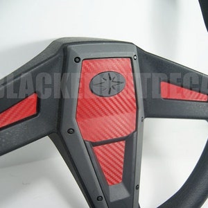 May include: Black steering wheel with red carbon fiber accents. The steering wheel has a star-shaped logo in the center.