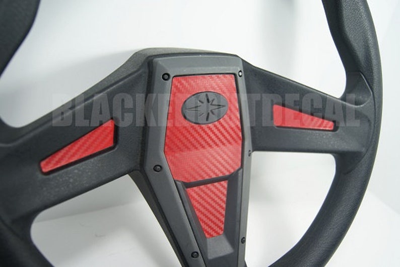 May include: Black steering wheel with red carbon fiber accents. The steering wheel has a star-shaped logo in the center.
