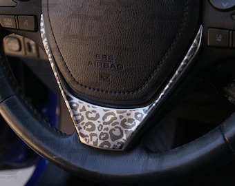 May include: Close-up of a car steering wheel with a black leather grip. The center trim features a silver leopard print design. The airbag is labeled "SRS AIRBAG".