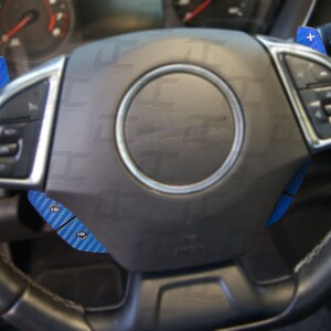 May include: A black leather steering wheel with blue carbon fiber paddle shifters. The steering wheel has a silver trim and a black center with a silver ring around it.