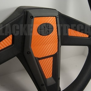 May include: A black steering wheel with orange carbon fiber accents. The steering wheel has a black center with a star-shaped logo.