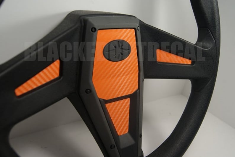 May include: A black steering wheel with orange carbon fiber accents. The steering wheel has a black center with a star-shaped logo.