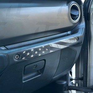 May include: A black and white distressed American flag decal on a black car interior door panel. The decal is placed on the door handle trim.