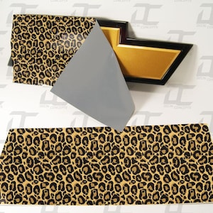 May include: A sheet of leopard print vinyl with a gold and black Chevy emblem. The vinyl is being applied to the emblem.