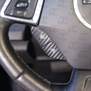 May include: Close-up of a black leather steering wheel with two black buttons labeled "FAV" on a carbon fiber panel.