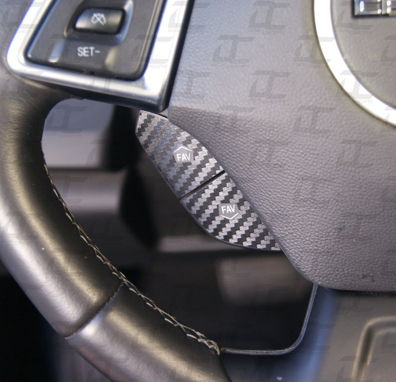 May include: Close-up of a black leather steering wheel with two black buttons labeled "FAV" on a carbon fiber panel.
