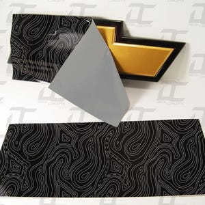 May include: A black and white topographic pattern decal for a car emblem. The decal is on a grey backing sheet and is shown with a gold and black car emblem.