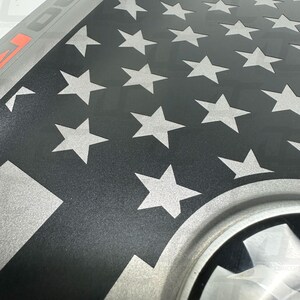 May include: A black and silver American flag decal with a white stripe and a silver star pattern. The decal is designed to be applied to a vehicle.