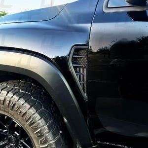 K&ouml;nnte beinhalten: Schwarze Fahrzeugkotfl&uuml;gel-Entl&uuml;ftung mit einem schwarz-wei&szlig;en Design der amerikanischen Flagge. Die Entl&uuml;ftung befindet sich an der Seite des Fahrzeugs, in der N&auml;he des Vorderrads. Der Reifen ist schwarz mit einem robusten Profil.