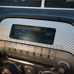 以下が含まれることがあります： 時間、ラジオ局、その他の情報を表示するデジタルディスプレイを備えた車のダッシュボード。ディスプレイは、ラジオとCDプレーヤーの機能のためのボタンとノブに囲まれています。ダッシュボードはカーボンファイバーパターンです。