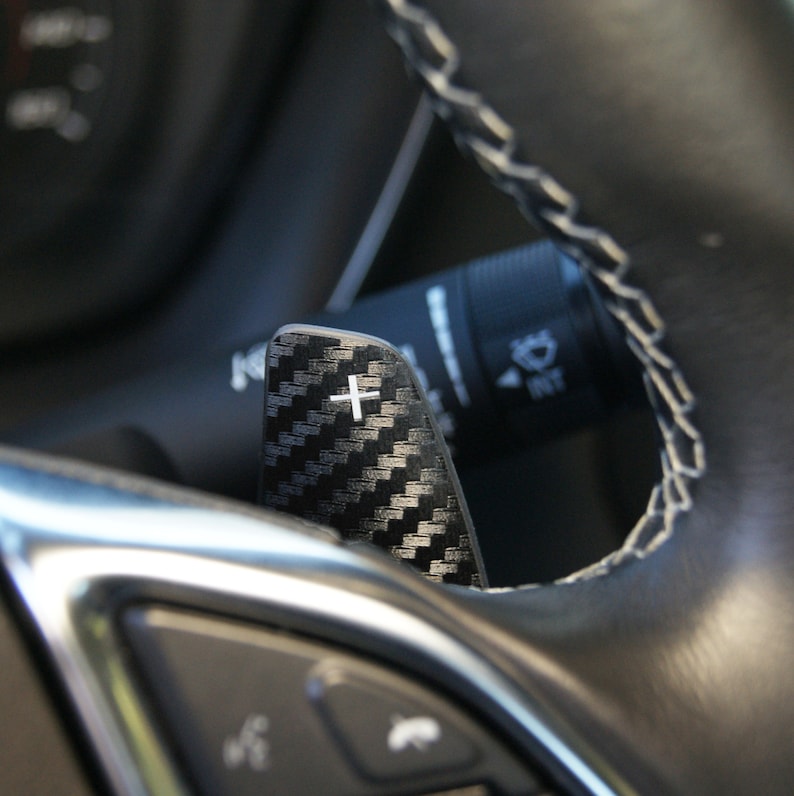 May include: Close-up of a black carbon fiber paddle shifter with a white plus sign. The paddle shifter is attached to a black leather steering wheel.