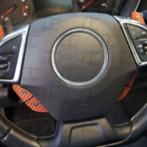 May include: A black leather steering wheel with orange carbon fiber accents. The steering wheel has buttons and controls on the left and right sides.