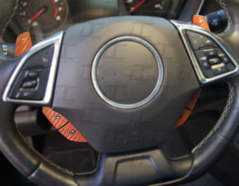 May include: A black leather steering wheel with orange carbon fiber accents. The steering wheel has buttons and controls on the left and right sides.