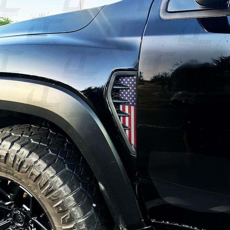 K&ouml;nnte beinhalten: Schwarzer Fahrzeugkotfl&uuml;gel mit einem Design der amerikanischen Flagge. Die L&uuml;ftung hat einen schwarzen Rahmen mit den Sternen und Streifen der Flagge. Reifen und Rad sind ebenfalls sichtbar.